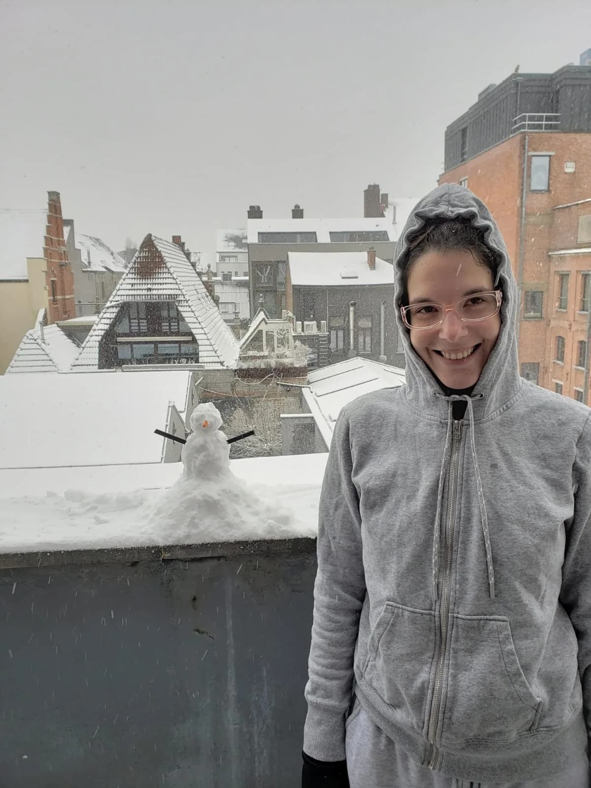 Laís num terraço com neve em Antuérpia, sorrindo ao lado de um bonequinho de neve